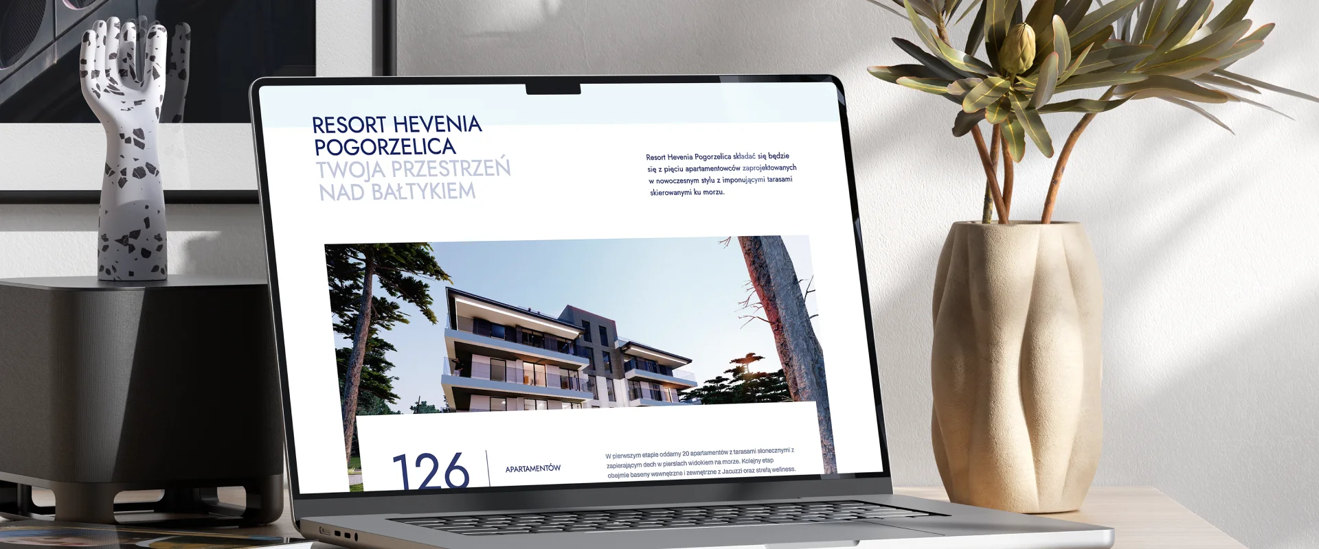 Obraz przedstawia wersję mobilną strony Hevenia, promującą nadmorskie apartamenty w Mrzeżynie. Na ekranie telefonu widoczna jest część strony z nazwą „Hevenia Mrzeżyno” oraz przyciskiem „Odnajdź swój apartament”. W tle widoczny jest nowoczesny budynek apartamentowy z przestronnymi balkonami.